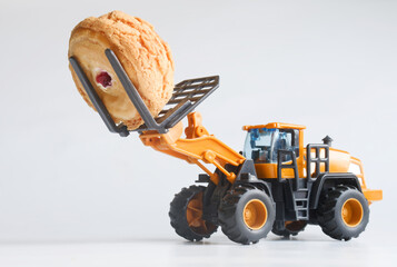 Yellow toy forklift lifts a fresh cream-filled cookie on white; playful bakery delivery and loading concept for confectionery ads, sweet and kid-friendly visual. Photo