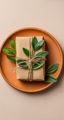 Gift wrapped in kraft paper, tied with twine, and decorated with small leaves, rests on a terracotta plate against a beige background
