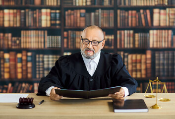 Judge in courtroom reads law documents carefully. At a wooden desk, gavel and scales near the robed...