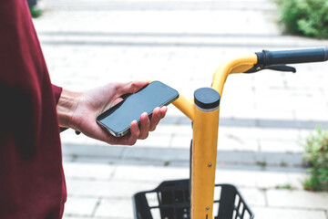 A person holds a smartphone next to the handlebar of a yellow rental bicycle on a city street, using a mobile app to unlock the bike for urban transportation and eco-friendly commuting