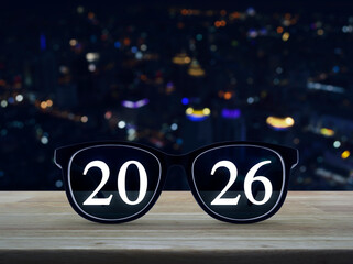 2026 white text on black eye glasses on wooden table over blur colorful night light modern city tower and skyscraper, Business vision happy new year 2026 concept