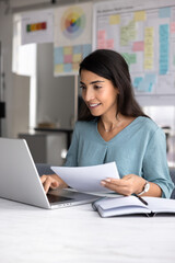 Positive Hispanic accountant woman checking electronic and paper financial reports