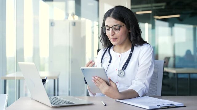 Confident young female doctor talking on video call using digital tablet in hospital clinic. Smiling medical worker physician is having remote consultation with patient sitting at workplace in office - Powered by Adobe