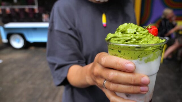 Young woman holding an iced matcha dalgona latte with whipped green tea foam and strawberry garnish in a plastic cup, refreshing summer street food market beverage