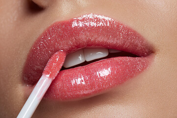 Naklejka premium Close-up of glossy pink lips of a young woman applying liquid lip gloss, showcasing beauty, makeup, cosmetics, fashion, and self-care concepts for consumer products