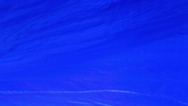 A bright blue textured surface is visible. The lighting highlights the subtle ripples and irregularities in the material. It resembles a close-up of synthetic fabric, plastic sheeting, or tarpaulin.