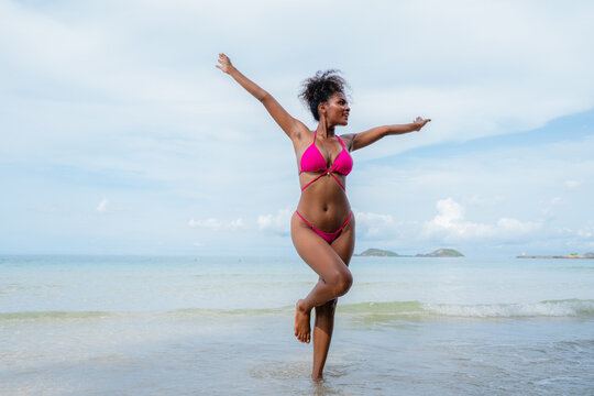 Beautiful African american woman in pink bikini on tropical beach. Portrait of dark skinned woman smiling at sea. Brunette tanned girl in swimwear enjoying and walking on beach.