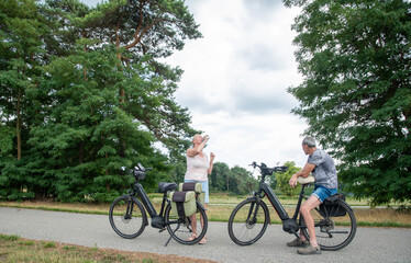 Obraz premium Elderly couple enjoys active retirement by riding modern electric bikes along scenic nature trails, while a woman drinks water and relaxes in the fresh air during summer vacation