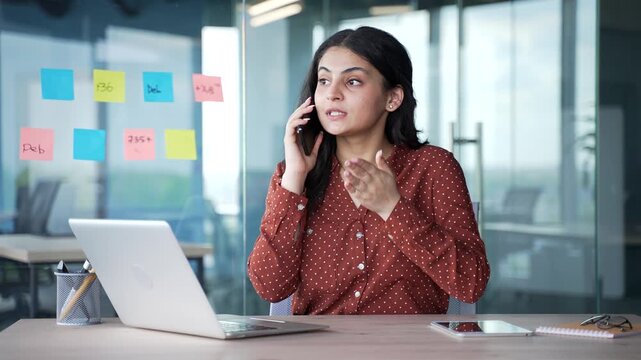 Confident young businesswoman talking on mobile phone while sitting at workplace in modern business office. Busy female employee discusses, communicates with partner or has a conversation with client