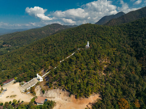 Aerial view of the gleaming white Big Buddha statue perched atop a verdant mountain amidst a tapestry of emerald trees, Big Buddha Pai, Mae Hi, Thailand.