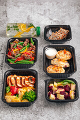 Healthy fitness meals in plastic containers with grilled vegetables, meat, snacks, and infused water on gray concrete background