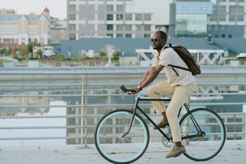 Young adult Black man riding bicycle along urban waterfront, wearing sunglasses and backpack, looking ahead with focused expression, modern city buildings reflected in water