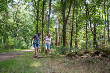 Senior couple enjoying an outdoor summer stroll, walking through a serene forest path, observing something interesting on the ground, engaging in active retirement and nature exploration