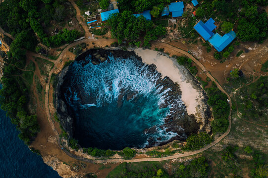 Aerial view of a striking natural pool with turquoise waters crashing against the sandy shore, surrounded by verdant cliffs, Nusa Penida, Bali, Indonesia.