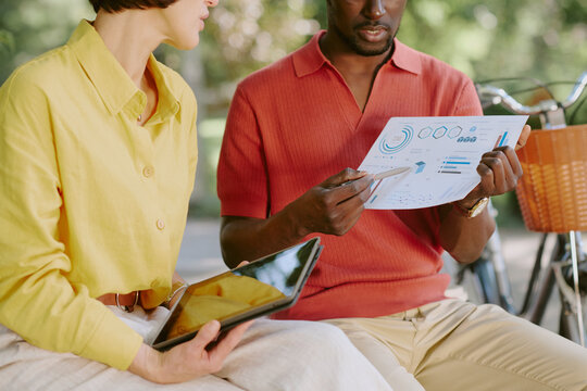 Caucasian middle aged woman and Black middle aged man discussing business analytics outdoors, holding digital tablet and paper chart with graphs, collaborating on project