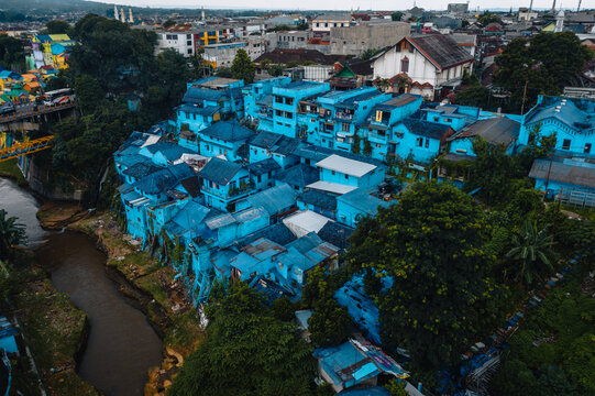 Aerial view of vividly painted blue buildings nestled amongst lush greenery, contrasting with the river and urban landscape, Jodipan Rainbow Village, Malang, Indonesia.