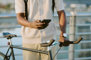 Young adult Black man standing outdoors holding smartphone in one hand and gripping bicycle handlebars with other hand, wearing wristwatch and backpack, using mobile device