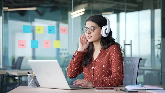 Happy young businesswoman in wireless headphones listens to music during break at work on a laptop sitting at workplace in business office. Joyful woman turns on playlist on computer and enjoying song