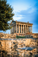 Vertical view of the Temple of Garni in Armenia with foreground ruins and a large tree. Captured at sunset, highlighting the temple?s ancient columns and golden tones