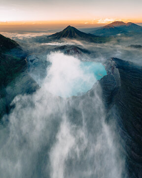Aerial view of the ethereal, turquoise crater lake shrouded in mist, with the soft sunrise casting a glow over the distant peaks, Mt. Ijen, Jawa Timur, Indonesia.