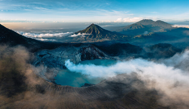 Aerial view of the stunning turquoise crater lake steaming amidst swirling clouds, framed by rugged peaks piercing the horizon, Mt. Ijen, Jawa Timur, Indonesia.