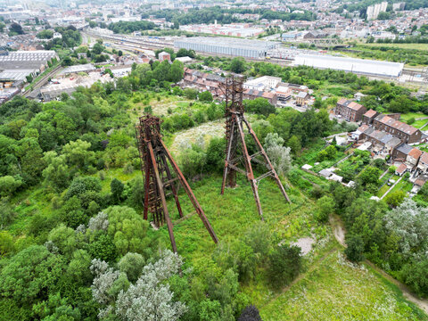 Aerial view of two weathered headframes standing as silent giants amidst vibrant greenery, a stark contrast to the industrial backdrop, Charleroi, Hainaut, Belgium.