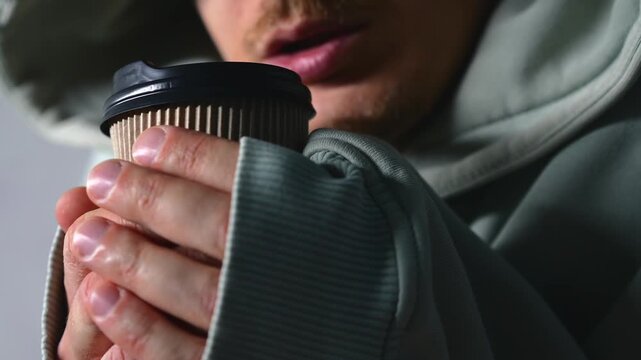 A frozen man in a warm hooded sweatshirt drinks a hot drink to warm up on a cold winter evening.
A warming drink in cold temperatures.
Warm up, drink, tea, frozen