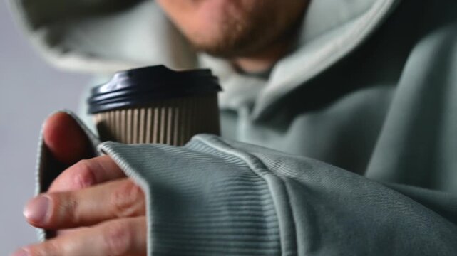 It's cold in the apartment and a man is drinking a hot drink to warm up.
A man is shivering from hypothermia indoors.
A guy in a thick hoodie is trying to warm up and drinking cold medicine.