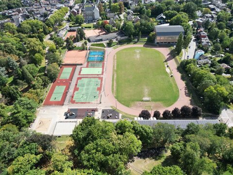 Aerial view of the vibrant sports complex with red tennis courts, blue basketball courts, and a bright green football field amidst lush trees, Li&Atilde;&uml;ge, Wallonia, Belgium.