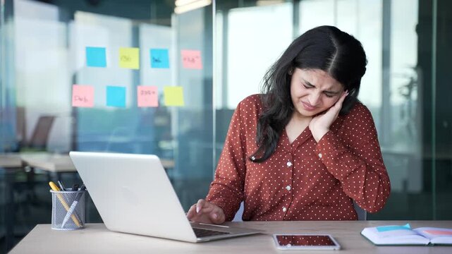 Young businesswoman suffers from ear pain while sitting at workplace in office. Upset woman touches her ear with her fingers. She has furuncle, shingles, eczema, ear diseases or otitis media. Earache