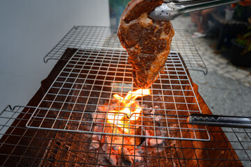 Grilled beef steak on barbecue grill over charcoal fire with tongs. Tender meat sizzling on a barbecue grill with flames below.