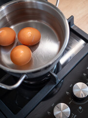 Three brown eggs in a stainless steel pot on a gas stove with water, preparation for boiling hard boiled eggs in a home kitchen.
