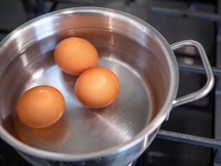 Three brown eggs in a stainless steel pot on a gas stove with water, preparation for boiling hard boiled eggs in a home kitchen.
