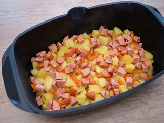 Boiled potatoes, carrots and sliced sausages layered in a black baking dish, prepared for traditional French-style baked potato casserole.