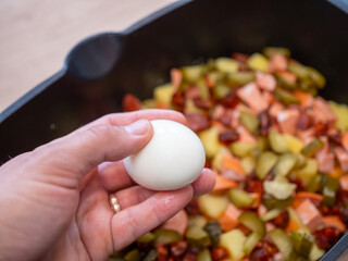 Adding boiled egg to layered dish with potatoes, carrots, sausage, chorizo, and pickles for homemade French potatoes casserole preparation.
