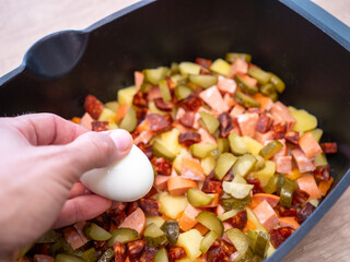 Adding boiled egg to layered dish with potatoes, carrots, sausage, chorizo, and pickles for homemade French potatoes casserole preparation.
