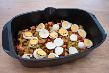 Preparation of traditional French potatoes with sausage, eggs, and pickles in a black baking dish before baking. Homemade Slovak comfort food.