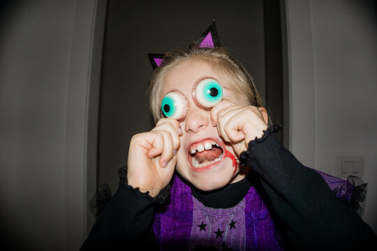 A young child in a Halloween costume with fake eyeballs and blood splatters on their face makes a funny, spooky expression.
