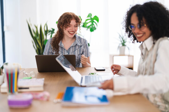 Two women collaborate at a desk, one with curly red hair laughing while using a laptop, the other with dark curly hair smiling and working on her own device.