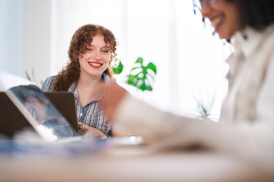 Two women collaborate on a project, one smiling as she looks at a laptop screen, the other focused on documents.