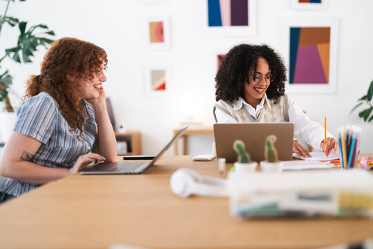 Two women collaborate at a wooden desk, one using a laptop and the other writing in a notebook, surrounded by art and plants.