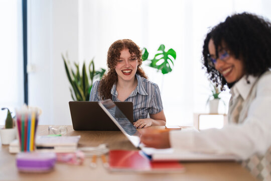 Two women collaborate at a desk with laptops and notebooks, one laughing while the other writes.