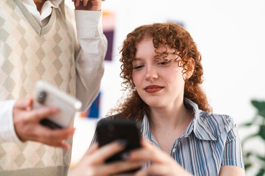 Two young women are looking at their phones, one with curly red hair and a nose ring, the other in a argyle sweater vest.