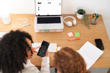Two women collaborate at a desk, one using a smartphone calendar and the other a notebook, surrounded by work essentials like a laptop and headphones.