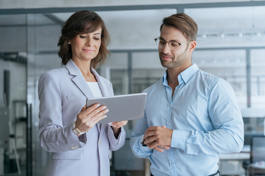 Mature Latin business woman and European young man discussing finance project on tablet gadget in office. Two diverse partners team, professional business people mentor and intern working together