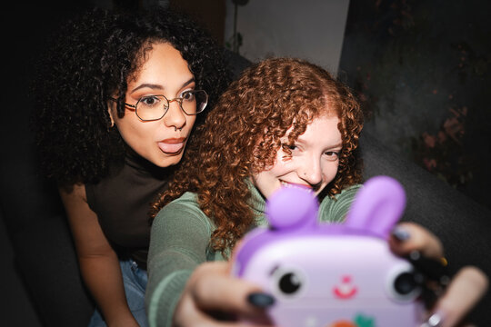 Two young women with curly hair take a selfie with a purple toy camera, one sticking her tongue out playfully.