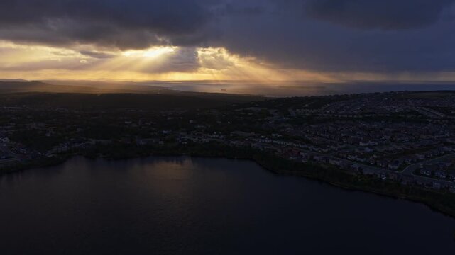 Aerial footage of Conception Bay at dusk where dramatic godrays pierce heavy clouds to illuminate dark waters, creating a moody yet serene atmosphere above Newfoundland.