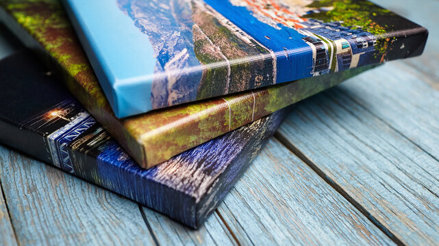 Three colorful canvas photo prints stacked on a rustic blue wooden surface, showing textured edges and vibrant printed landscapes