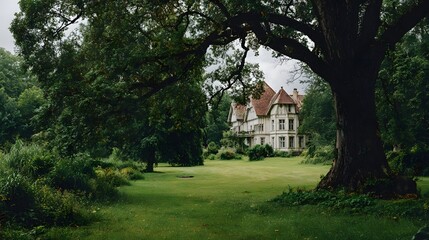 Fototapeta premium Majestic old tree frames a beautiful, grand dwelling on a sprawling green lawn; lush greenery surrounds the elegant residence.