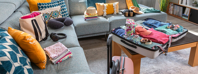 Banner of suitcase packed with clothes and accessories for summer vacation placed next to sofa in the living room, suggesting an upcoming trip. Open suitcase with colorful clothes ready for vacation.
